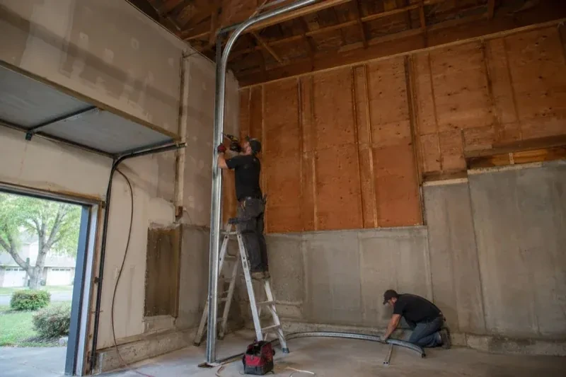 One person on a stepladder working beside a tall curved garage-door track while another kneels beside loose track sections in an unfinished garage. — High-Lift Garage Door Conversion, Brampton