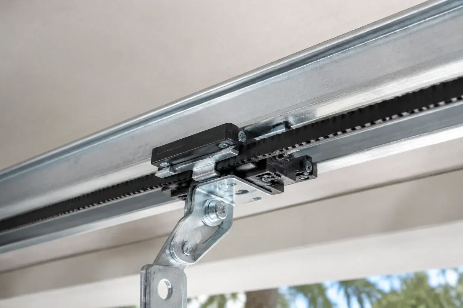 Black plastic trolley and bent galvanized bracket on a steel opener rail with a toothed black belt, against a ceiling. — Belt-Drive Opener Repair, Brampton